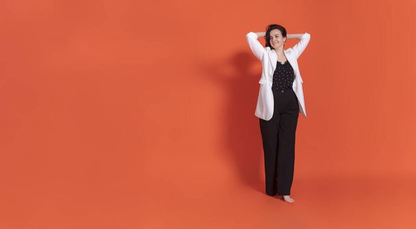 Young brunette woman in black trouser suit and white jacket stands on orange background. Raised both hands and launched them into their hair. - Photo, image