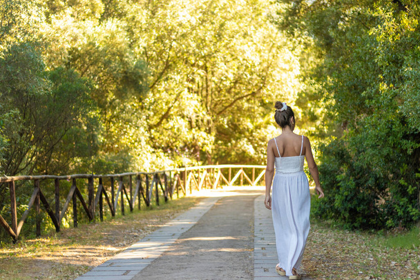 Eine unkenntlich zurückgebliebene junge brünette Frau, die im weißen Sommeroutfit zur Seite schaut und auf einem leeren Pfad durch die Natur geht - Foto, Bild