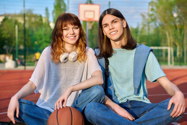 Portrait of couple friends teenagers guy and girl looking at camera, sitting on school basketball court. Teenage students with backpacks, ball in sunset. Active healthy sport lifestyle, youth concept - Fotografie, Obrázek