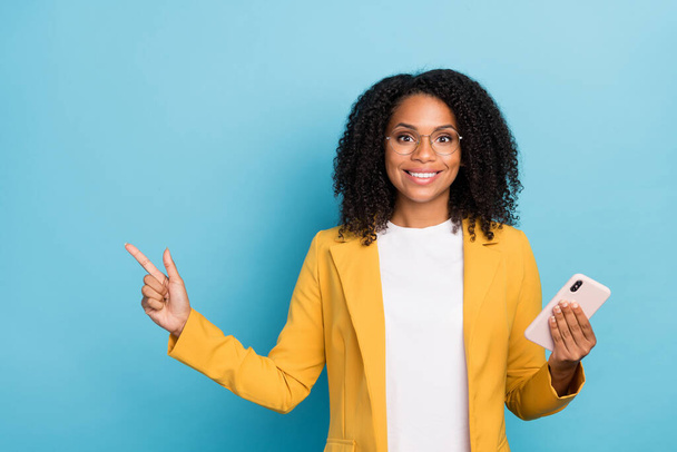 Photo of young black woman happy positive smile hold mobile point finger empty space ad promo isolated over blue color background. - 写真・画像