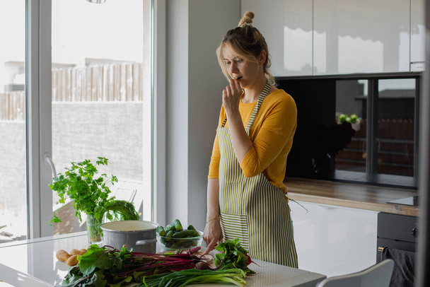 Önlüklü kadın salatalık tadıyor, mutfakta yemek yapmak için sebze fişi seçiyor. Öğlen yemeği için taze organik salatalık, patates ve pancar. Tatillerde diyet - Fotoğraf, Görsel