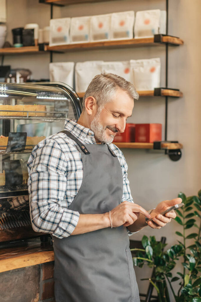 Message. Smiling gray-haired man in apron touching smartphone screen standing sideways to camera with back to showcase in cafe - Foto, Bild