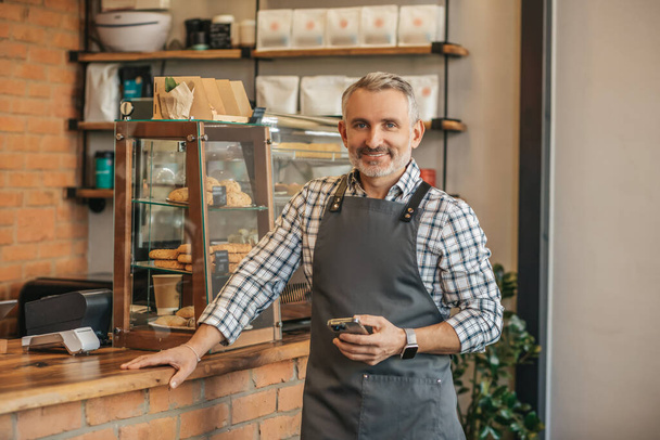 Small business. Confident gray-haired man in apron holding smartphone leaning with hand on bar counter standing smiling at camera in cafe - Valokuva, kuva
