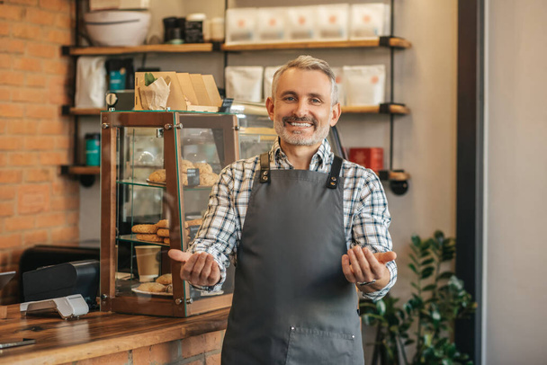 Welcome. Friendly middle-aged man in apron standing in cafe near counter with desserts gesturing invitingly smiling at camera - Foto, imagen
