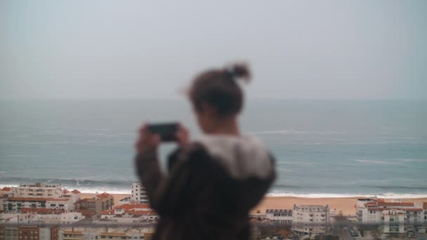 Teen boy scattare foto dal cellulare di una località turistica dal balcone dell'hotel e poi godersi una scena incredibile con grandi onde oceaniche rotolando sulla spiaggia, concentrarsi sullo sfondo. Turismo a Nazare, Portogallo - Filmati, video