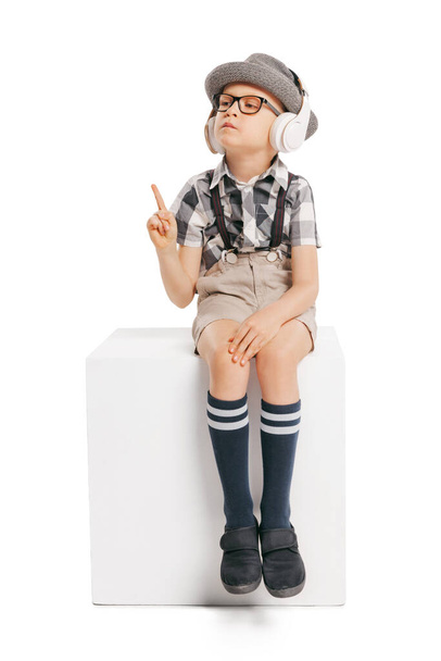 Listening to audio lesson. Cute little boy, serious kid in eyeglasses and hat isolated on white studio background with copyspace for ad. Concept of child emotions, facial expression, studying and ad. - Photo, Image