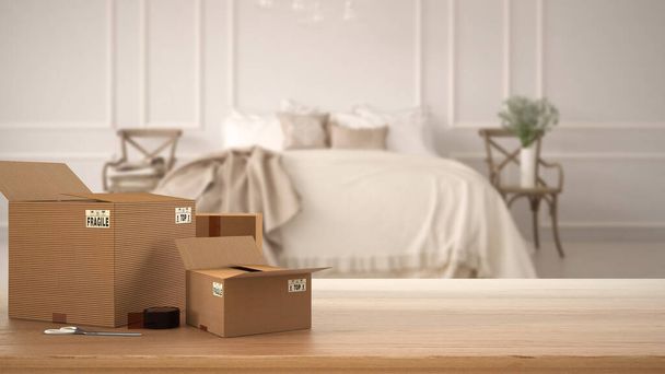 Wooden table, desk or shelf with stack of cardboard boxes over blurred view of vintage bedroom with soft bed and chair, classic interior design, moving house concept with copy space - Photo, Image