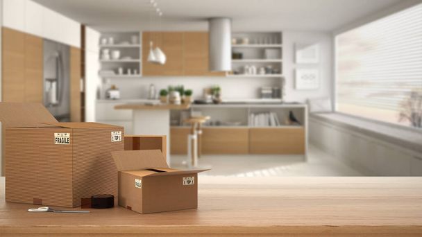 Wooden table, desk or shelf with stack of cardboard boxes over blurred view of minimalist kitchen with island, modern interior design, moving house concept with copy space - Photo, Image