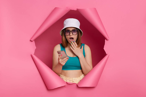 Portrait of shocked woman wearing tank top and panama posing in torn pink paper wall, using cell phone, reading shocking news, keeps mouth open covered with palm. - Foto, Imagen