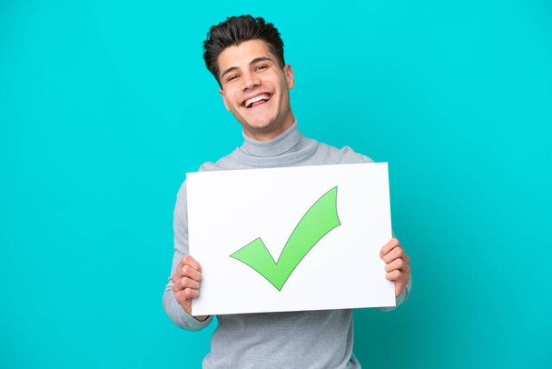 Young handsome caucasian man isolated on blue bakcground holding a placard with text Green check mark icon with happy expression - Photo, Image