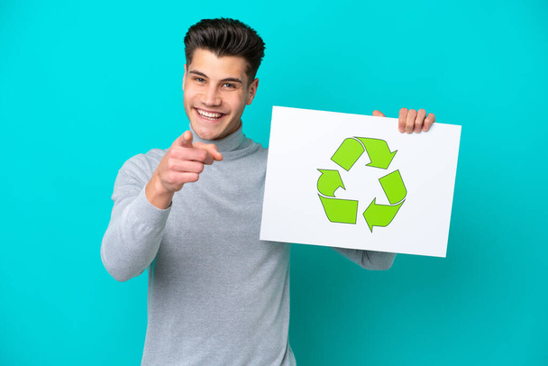 Junger gutaussehender kaukasischer Mann isoliert auf blauem Grund mit einem Plakat mit Recycling-Symbol und zeigt nach vorne - Foto, Bild