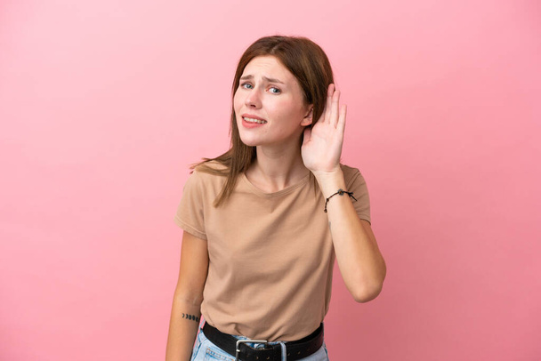 Νεαρή Αγγλίδα απομονωμένη σε ροζ φόντο ακούγοντας κάτι βάζοντας το χέρι στο αυτί - Φωτογραφία, εικόνα