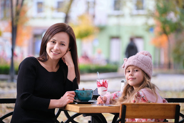 Eine junge hübsche Frau amüsiert sich an einem sonnigen Herbsttag mit ihrer kleinen Tochter im Straßencafé mit heißen Getränken. Glück in familiären Beziehungen. - Foto, Bild