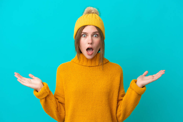 Молода англійська жінка, одягнена в зимовий піджак, ізольована на синьому фоні з шокованим виразом обличчя. - Фото, зображення