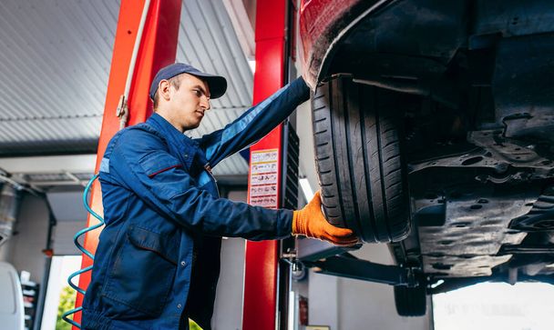 Expert specialist technician changes tires, tyres of lifted up car at auto service, wears uniform costume. - Photo, Image