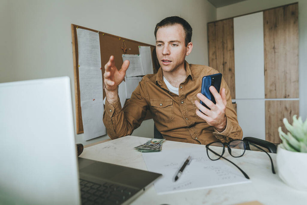 Молодий чоловік користується смартфоном і ноутбуком для відеобесіди, сидячи за столом у кімнаті. Freelancer проводить онлайн-конференцію. Використання технологій вдома. Вибіркове зосередження. - Фото, зображення