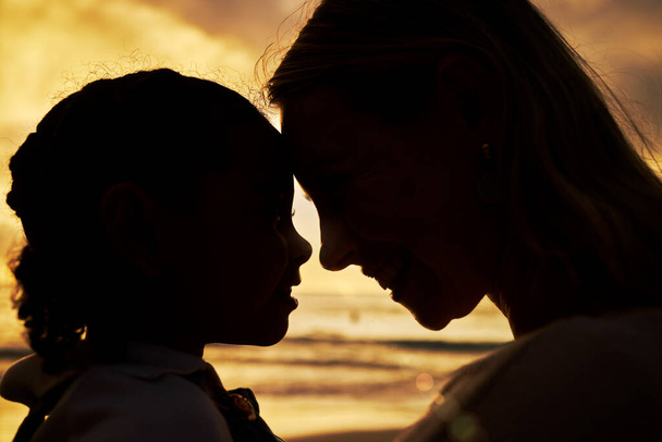 Blízká silueta matky a dcery, kteří při západu slunce stojí tváří v tvář na pláži. Podsvícená mladá žena a její dívka dítě s úsměvem a tváří k sobě s oceánem v pozadí. - Fotografie, Obrázek
