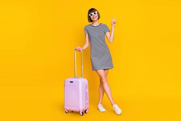 Full lenght photo of cool brunette young lady stand show v-sign with bag wear dress sneakers eyewear isolated on yellow background. - Valokuva, kuva
