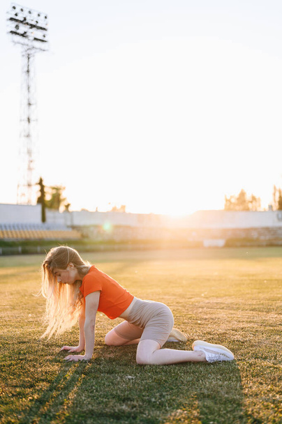 Pystysuora muotokuva nuori nainen urheiluvaatteissa tekee Cross Stretch on Grass. Tyttö treenaa illalla. Tyttö etuvalossa. Kultainen aika kesällä. Urheilija Golden Rays of Sun. - Valokuva, kuva