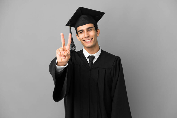 Joven graduada universitaria argentina aislada sobre fondo gris sonriendo y mostrando signo de victoria - Foto, imagen