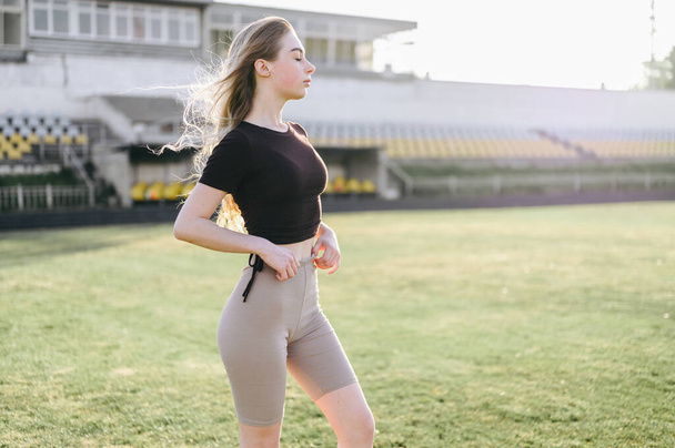 Young Woman in Black Top Adjusting Bicycles. Girl Stands Sideways Against Background of Sports Field. Practice on Summer Evening at Stadium - Photo, Image