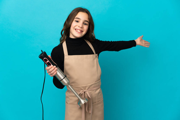Bambina utilizzando frullatore a mano isolato su sfondo blu che estende le mani di lato per invitare a venire - Foto, immagini