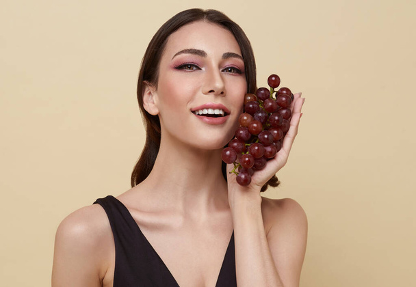 smiling woman with grape. Photo of beautiful woman with perfect makeup on nude color background. Beauty and Skin care concept. - Photo, Image