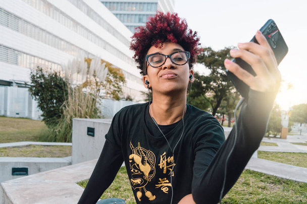 gyönyörű barna kolumbiai latin nő piros afro, szemüveg és jó hozzáállás ül a parkban naplementekor, hogy egy szelfit a telefonjával és üzleti épületek mögötte. - Fotó, kép