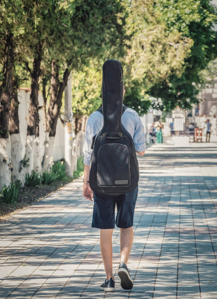 Πίσω όψη ενός άντρα που περπατάει με μια θήκη κιθάρας στην πλάτη του. - Φωτογραφία, εικόνα
