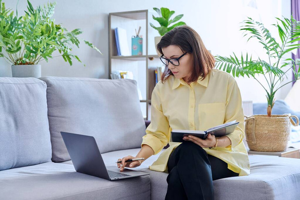 Mature businesswoman working on couch, using laptop making notes in business notebook. Confident middle aged female working from home office remotely. Business job freelancing, technology, 40s people - Foto, Imagem