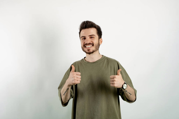 Vrolijke jongeman in groen t-shirt poserend geïsoleerd over een witte achtergrond glimlachend en met duimen omhoog voor de camera. Goedkeurende uitdrukking die naar de camera kijkt en succes laat zien. - Foto, afbeelding