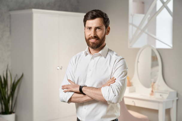 Handsome male businessman wearing white shirt posing, standing with crossed arms indoors. Front view of smiling entrepreneur looking at camera, with blurred bedroom on background. Concept of business. - 写真・画像
