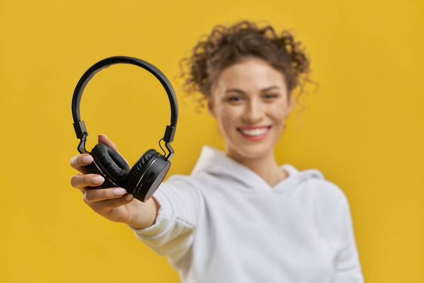 Neat female hand holding up black headphones indoor. Close up view of earphones supported by girls hand, with blurred figure of smiling woman, isolated on orange studio background. Concept of music. - Foto, immagini