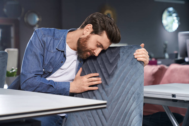 Middle-aged elegant bearded man leaning to chair back, while touching pattern before buying furniture. Gentle brunet male with closed eyes hugging soft grey chair in store. Concept of interior design. - Foto, Imagem