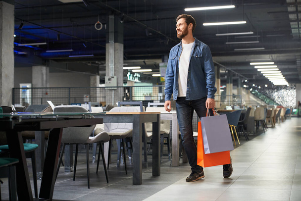 Handsome male shopper in casual outfit carrying paper bags, while walking in shopping mall. Front view of attractive guy with purchases strolling in furniture section of shop. Concept of man shopping. - Foto, Imagen