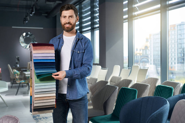 Confident male expert demonstrating various samples of upholstery fabric in studio. Front view of smiling bearded man showing colorful palette, with sunlit window on background. Concept of workplace. - Photo, image