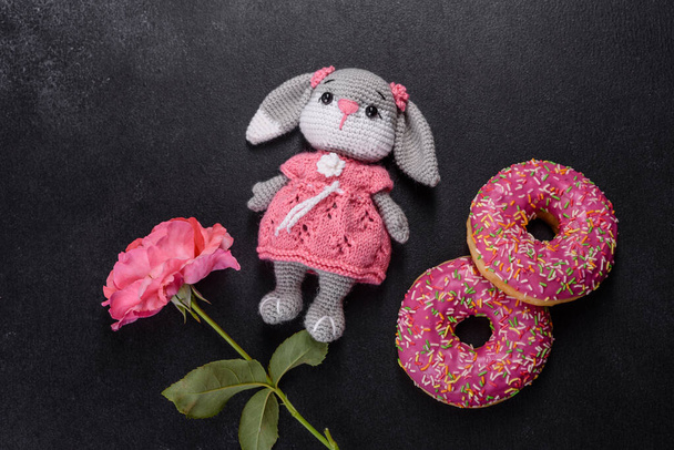 Güzel örülmüş tavşan oyuncağı, donutlı pembe jöleli ve koyu beton üzerine renkli serpiştirilmiş. - Fotoğraf, Görsel