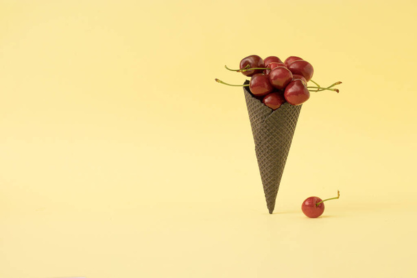 Trendy summer composition with fresh red cherries and black ice cream cone on pastel yellow background. Minimal summer food idea. - Foto, imagen