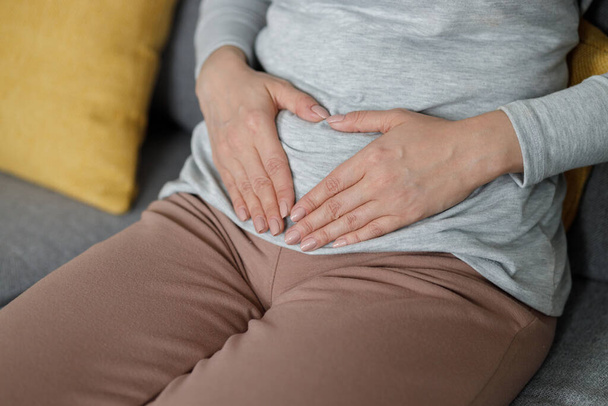 Sick young woman suffering from menstrual pain. Woman with hands squeezing belly having painful stomach ache or period cramps sitting on sofa, Abdominal pain, gastritis, and painful periods concept. - 写真・画像
