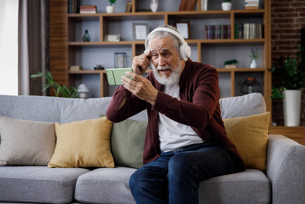 Happy handsome elderly gray-haired man in white headphones holding smartphone and laughing. Old man sitting on couch at home watching funny content videos blogs, TV shows, films. - Foto, immagini