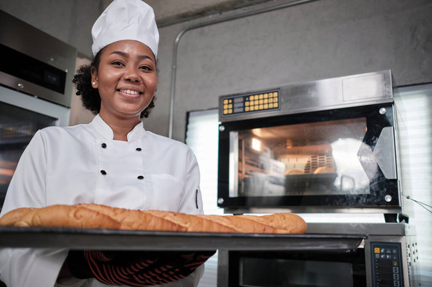Retrato de chef feminino afro-americano em uniforme de cozinha branca olhando para a câmera com sorriso alegre e orgulhoso com bandeja de baguete na cozinha, alimentos de pastelaria profissional, ocupação de padaria fresca. - Foto, Imagem