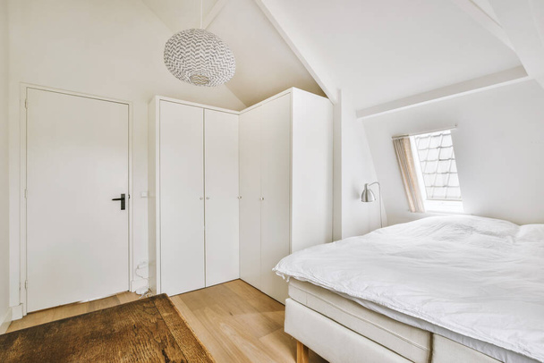 Moderní bytový interiér malé podkrovní ložnice s bílými stěnami, oknem a měkkou útulnou postelí a bílými skříněmi - Fotografie, Obrázek