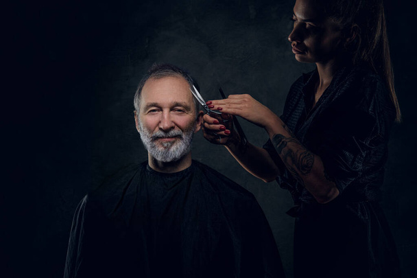 Retrato de barbeiro feminino cortando cabelos de seu cliente idoso alegre contra fundo escuro. - Foto, Imagem