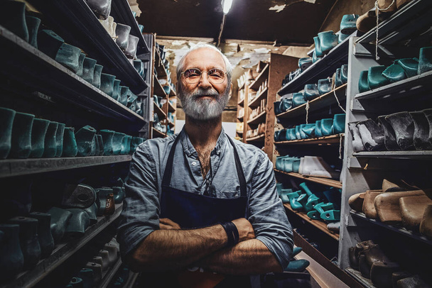 Portrait of senior man dressed in apron with crossed arms around shelfs with shoes. - Photo, image