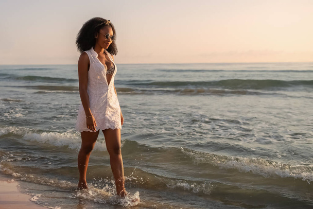 Afrikkalainen nainen, jolla on aurinkolasit, kesäinen valkoinen mekko ja bikinit kävelemässä rannalla auringonlaskun aikaan. Valikoiva painopiste. - Valokuva, kuva