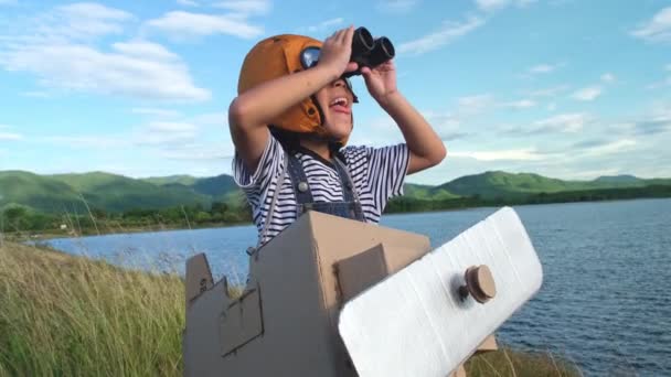 Linda niña soñadora jugando con aviones de cartón y binoculares en un prado junto al lago en un día soleado. Concepto de imaginación de sueño infantil. - Metraje, vídeo