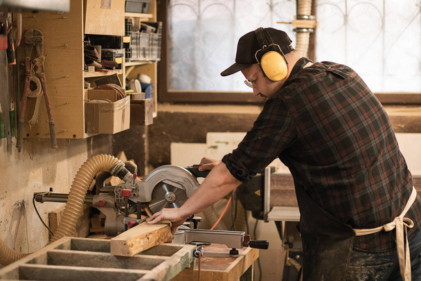 Joiner εργαζόμενος, επιχειρηματίας σε προστατευτική ποδιά και ακουστικά χρησιμοποιούν σύγχρονη μηχανή μύλος για την επεξεργασία κοπή σανίδων ξύλου, ξυλεία με κυκλικό πριόνι εργαστήριο, ξυλουργική απασχόληση. Οικοδομική βιομηχανία - Φωτογραφία, εικόνα