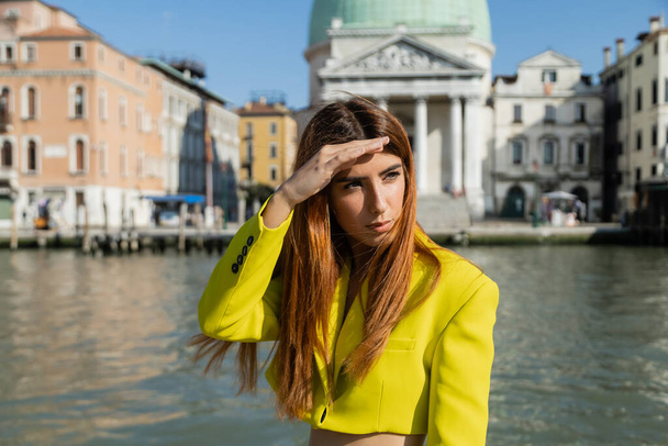 modische Frau, die die Hand über den Augen hält, während sie vor verschwommenem Hintergrund in Venedig wegschaut - Foto, Bild
