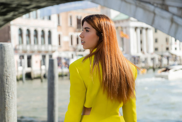 κοκκινομάλλα γυναίκα με κομψά κίτρινα ρούχα κοιτάζοντας μακριά στη Βενετία σε θολή φόντο - Φωτογραφία, εικόνα