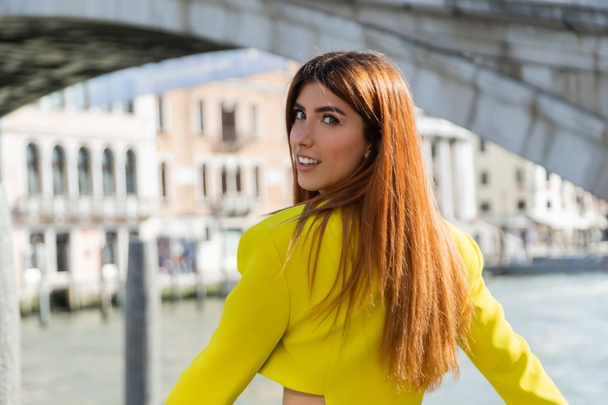 mujer pelirroja feliz en chaqueta de cosecha amarilla sonriendo a la cámara sobre fondo borroso en Venecia - Foto, Imagen